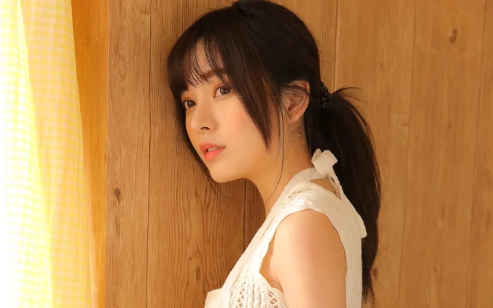 一名扎着马尾的女性穿着白色无袖上衣，站在木质墙面前。