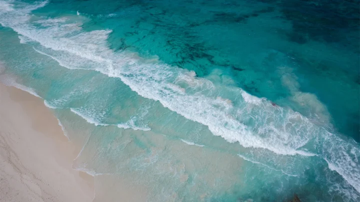 俯瞰视角下，碧 turquoise 海水中的白色浪潮拍打着浅色沙滩。