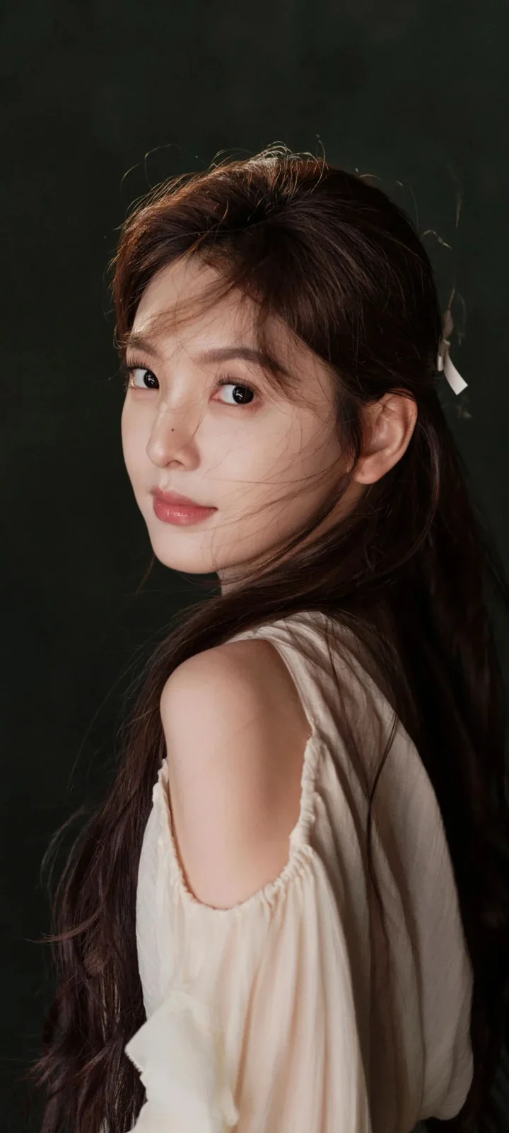긴 머리의 여성이 어두운 배경을 뒤로하고 크림색 오프숄더 상의를 입은 채 돌아보며 카메라를 바라보고 있다.