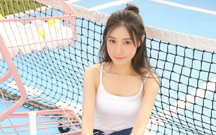 성인 여성이 파란 코트와 분홍색 금속 프레임을 배경으로 스포츠 코트의 네트 옆에 앉아 있습니다.