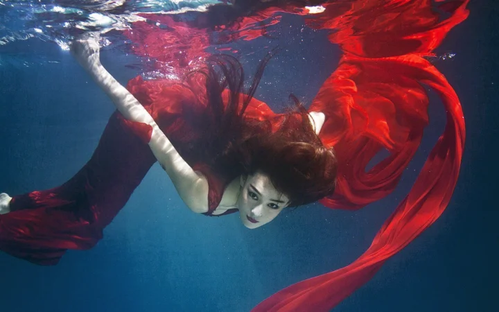 赤いドレスを着た成人女性が水中を泳ぎ、ドレスの裾と長い髪が水流に広がっています。