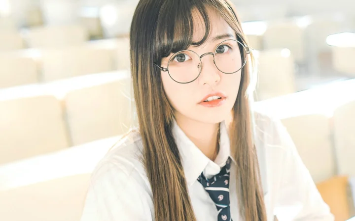 一位戴圆框眼镜、留长发的女性在明亮的室内环境中面向镜头。