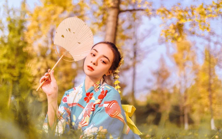 一名成年女子身穿蓝色和服，在秋日树林中手持折扇。