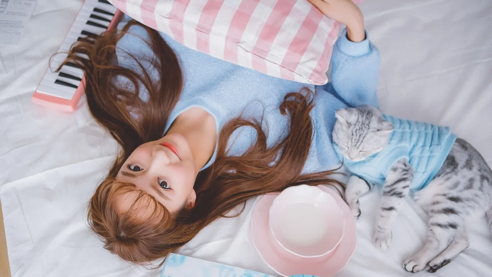 한 소녀가 침대에 누워 있고 파란 옷을 입은 고양이가 곁에 있으며 주변에 키보드와 쿠션, 그릇이 놓여 있습니다.