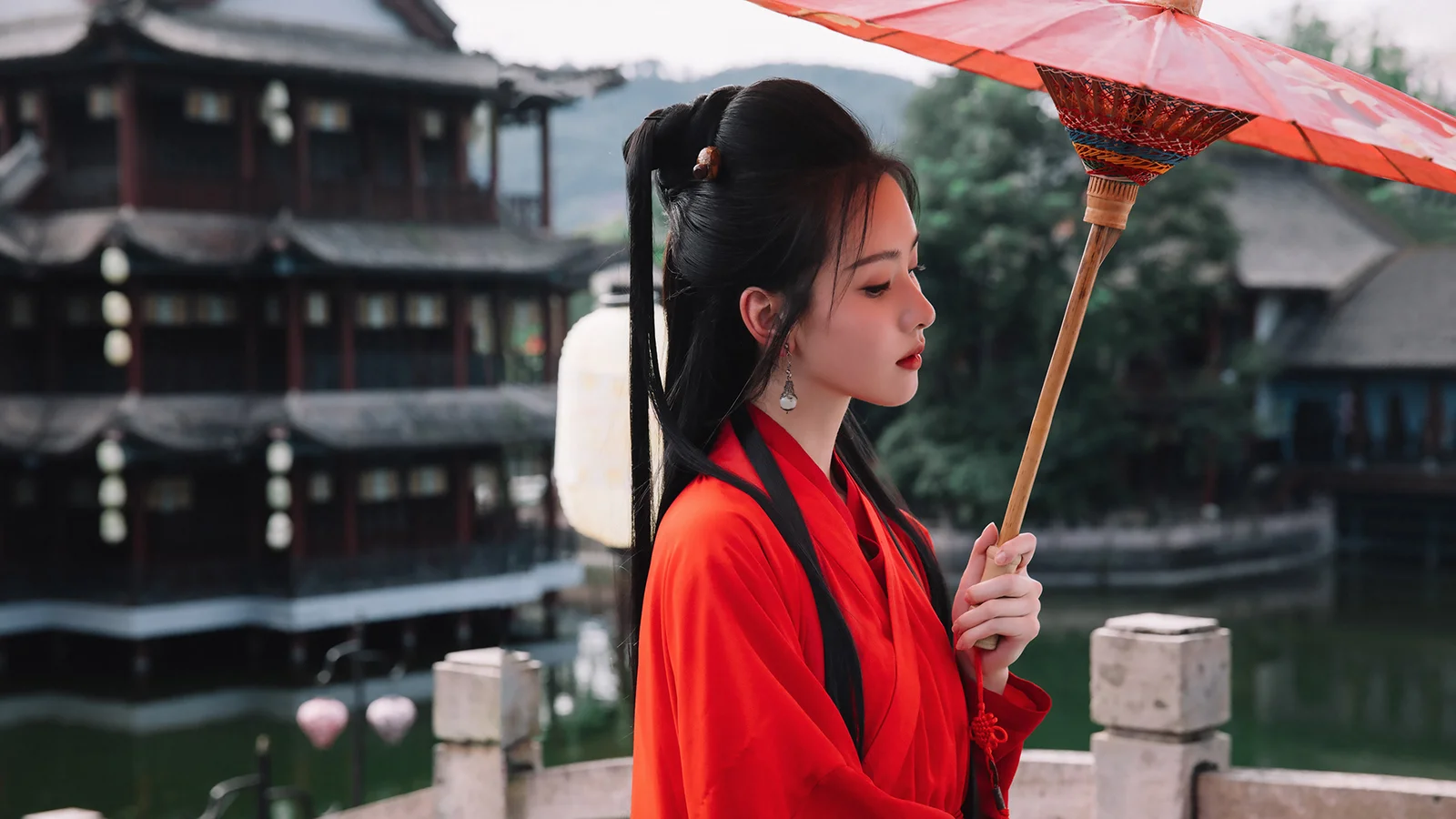 붉은 예복을 입은 여성이 전통적인 수변 건물 곁에서 붉은 양산을 들고 있다.