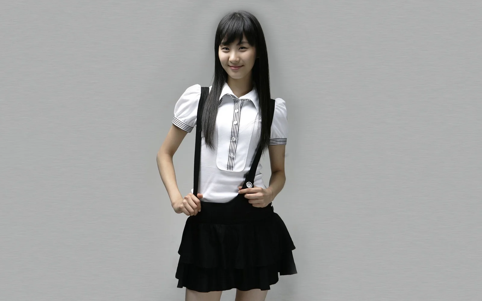 長い黒髪の女性が、白いトップスと黒いスカート姿でグレーの背景の前に立っています。