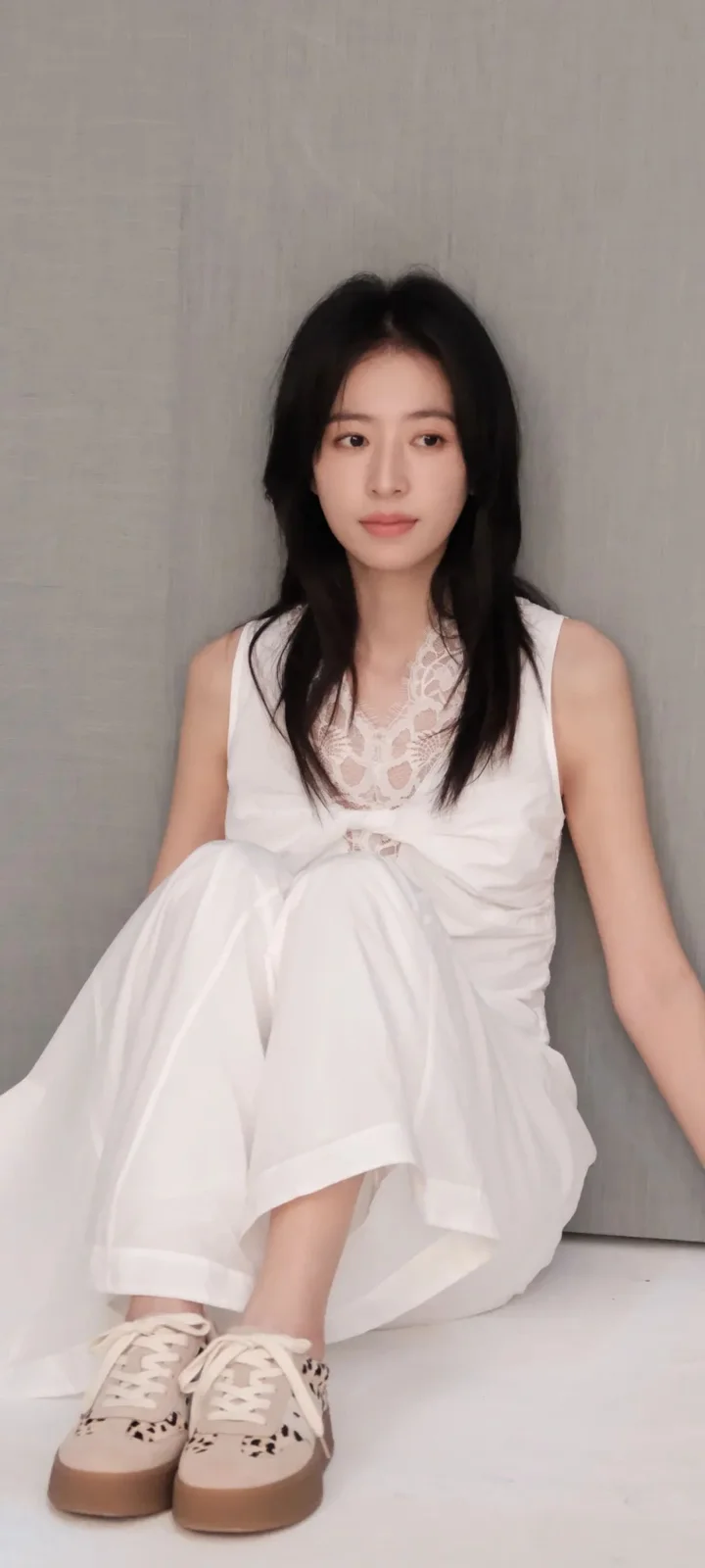 一名女子穿着白色连衣裙坐在室内墙边，脚穿运动鞋。