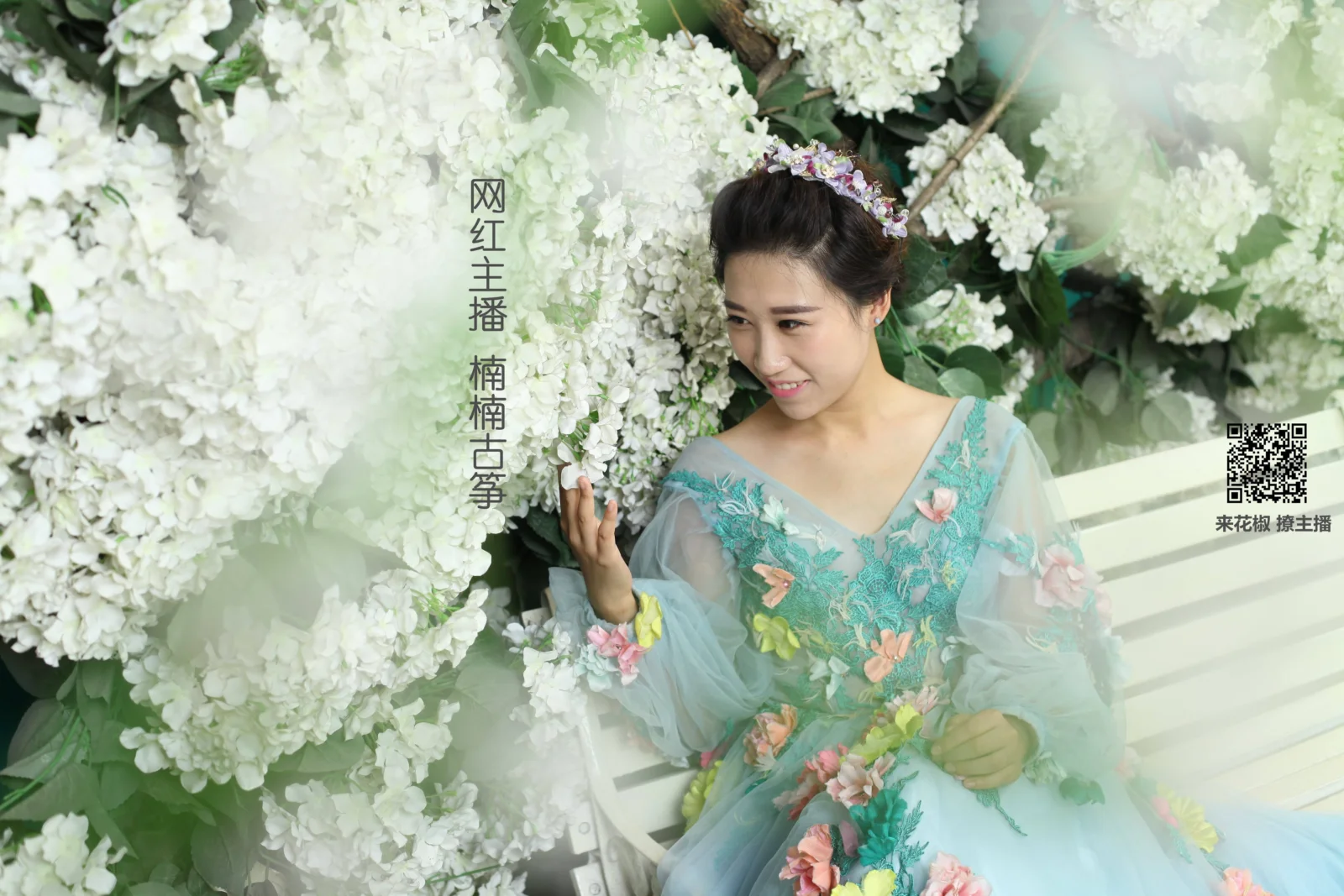 ターコイズ色の花柄ドレスを着た成人女性が、白い花に囲まれたベンチに座っています。