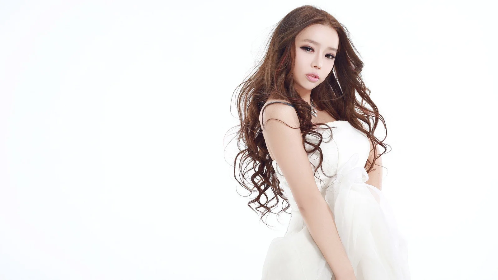 白いドレスを着た長髪の女性が白い背景の前に立っている。