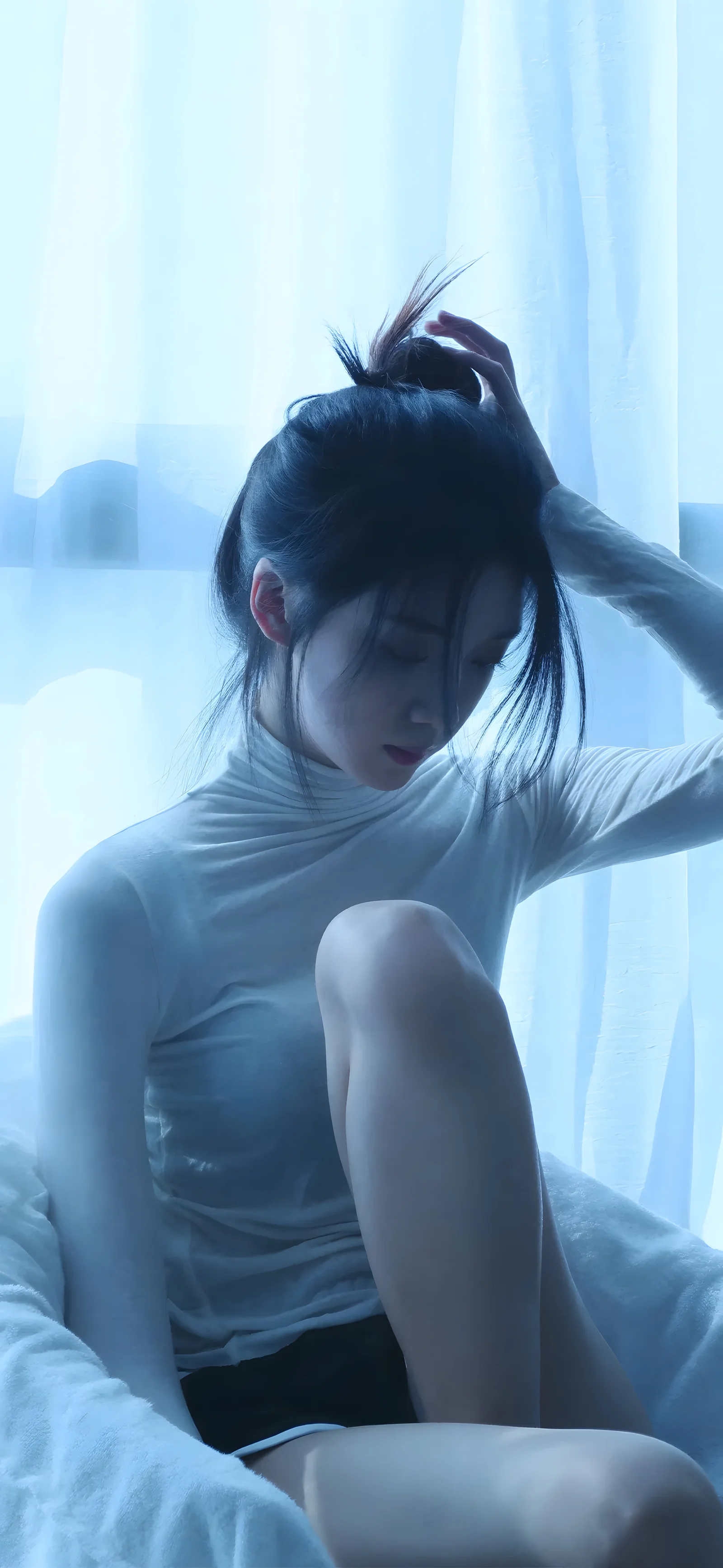 一名女子坐在柔和的蓝色光线中整理头发，身后是透光窗帘。