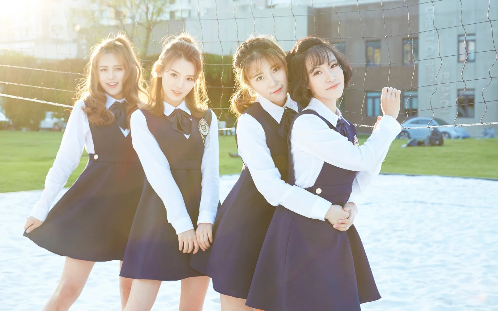 어두운 색상의 교복을 입은 네 명의 여성이 야외 배구 네트 앞에 서서 뒤쪽에서 비치는 햇빛을 받고 있습니다.