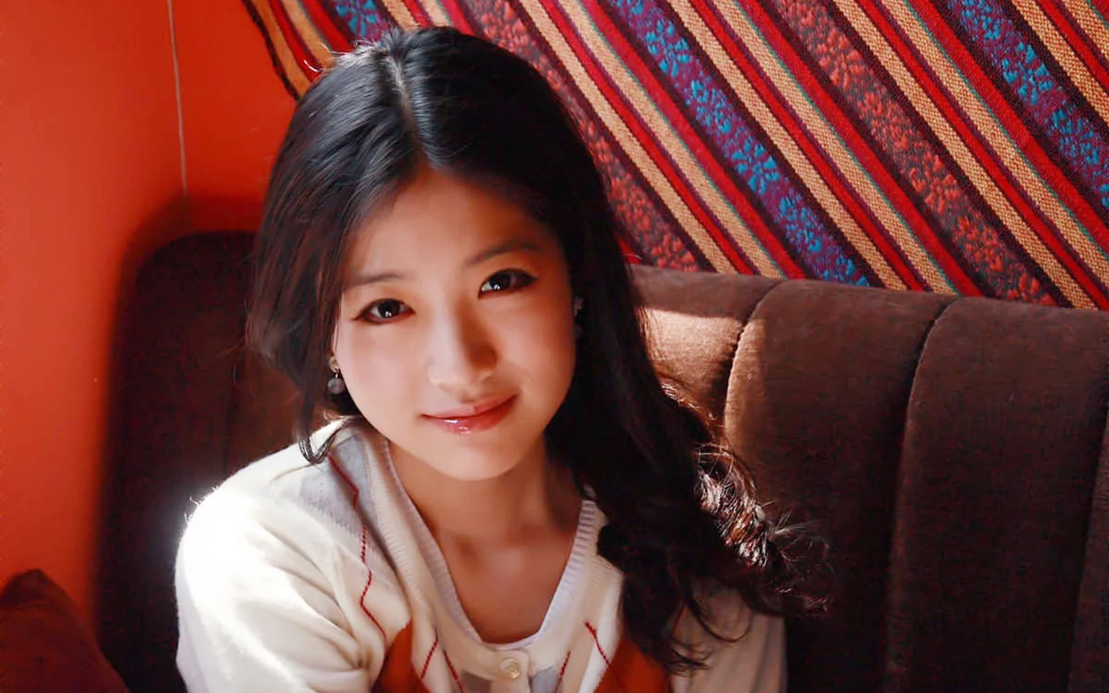 성인 여성이 실내 소파 앞에 앉아 뒤쪽의 화려한 줄무늬 천을 배경으로 미소 짓고 있다.