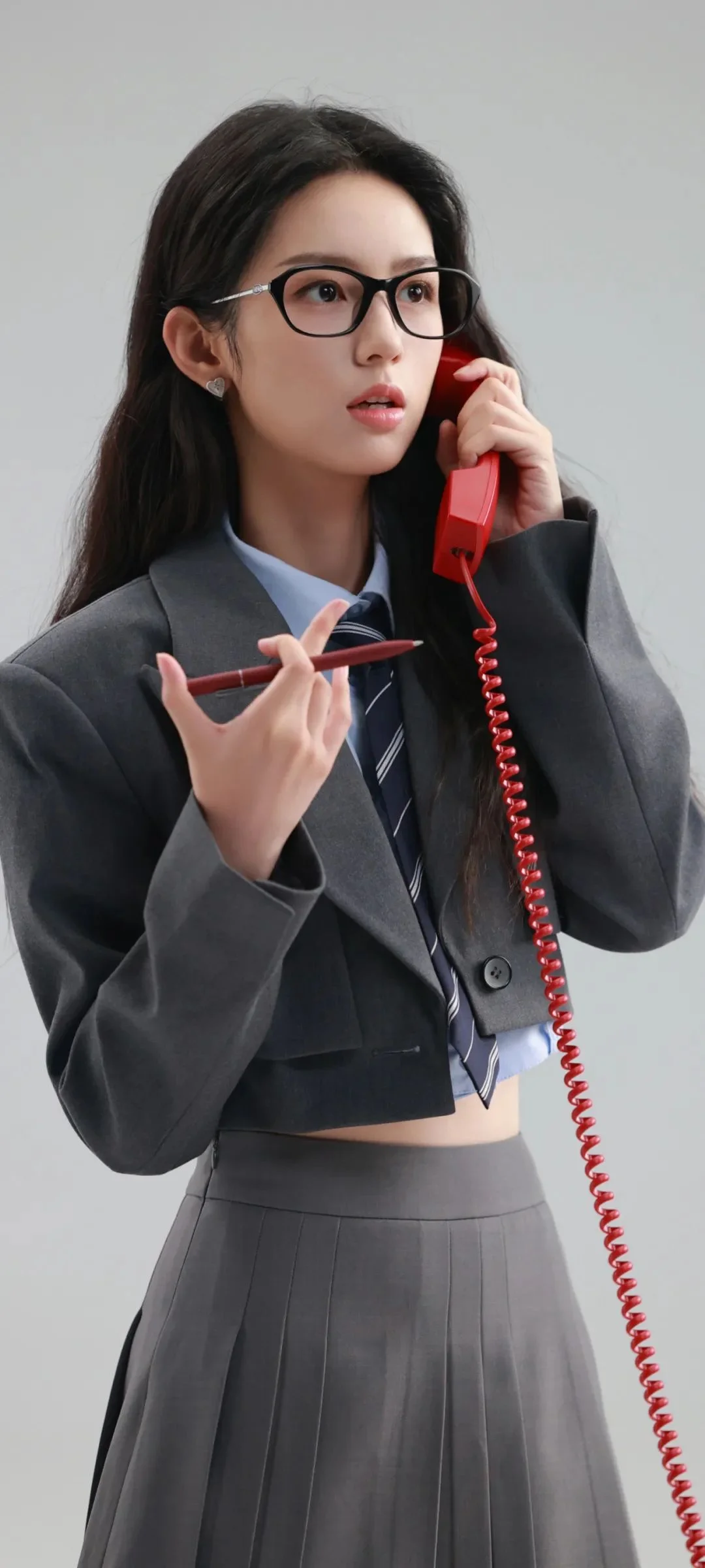 안경을 쓰고 회색 정장을 입은 여성이 빨간 전화 수화기와 스마트폰을 들고 있습니다.