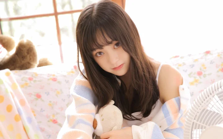긴 머리의 여성이 밝고 부드러운 실내에서 침대에 앉아 봉제인형을 안고 있습니다.