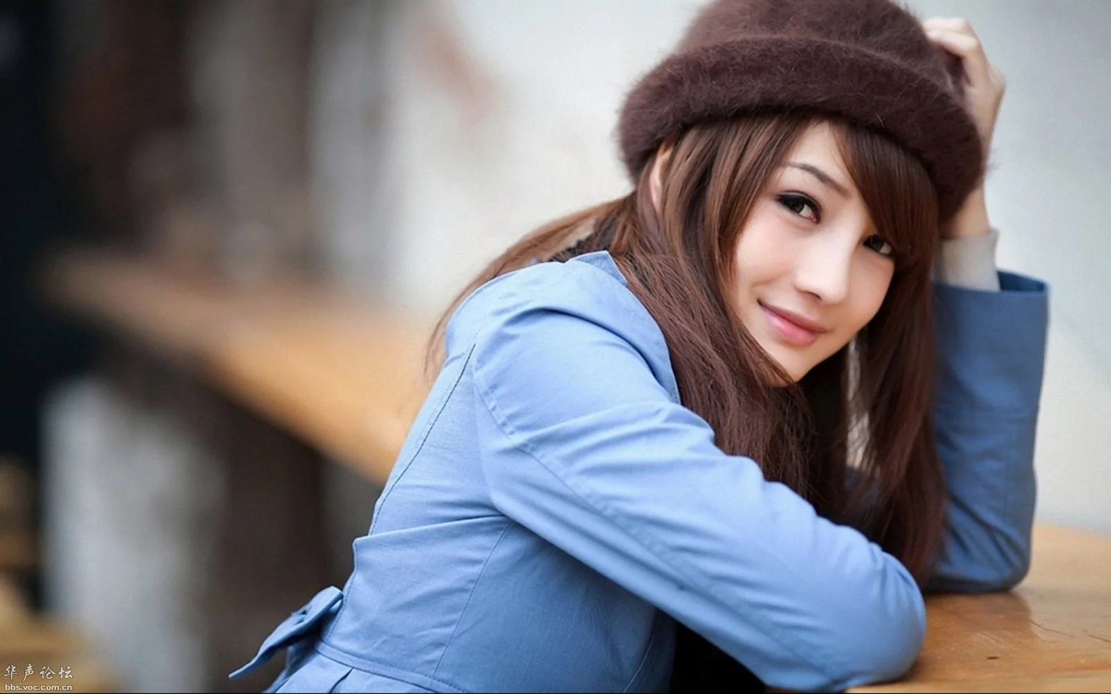 茶色の帽子と青いジャケットを身につけた女性が、テーブルに寄りかかってカメラを見ている。