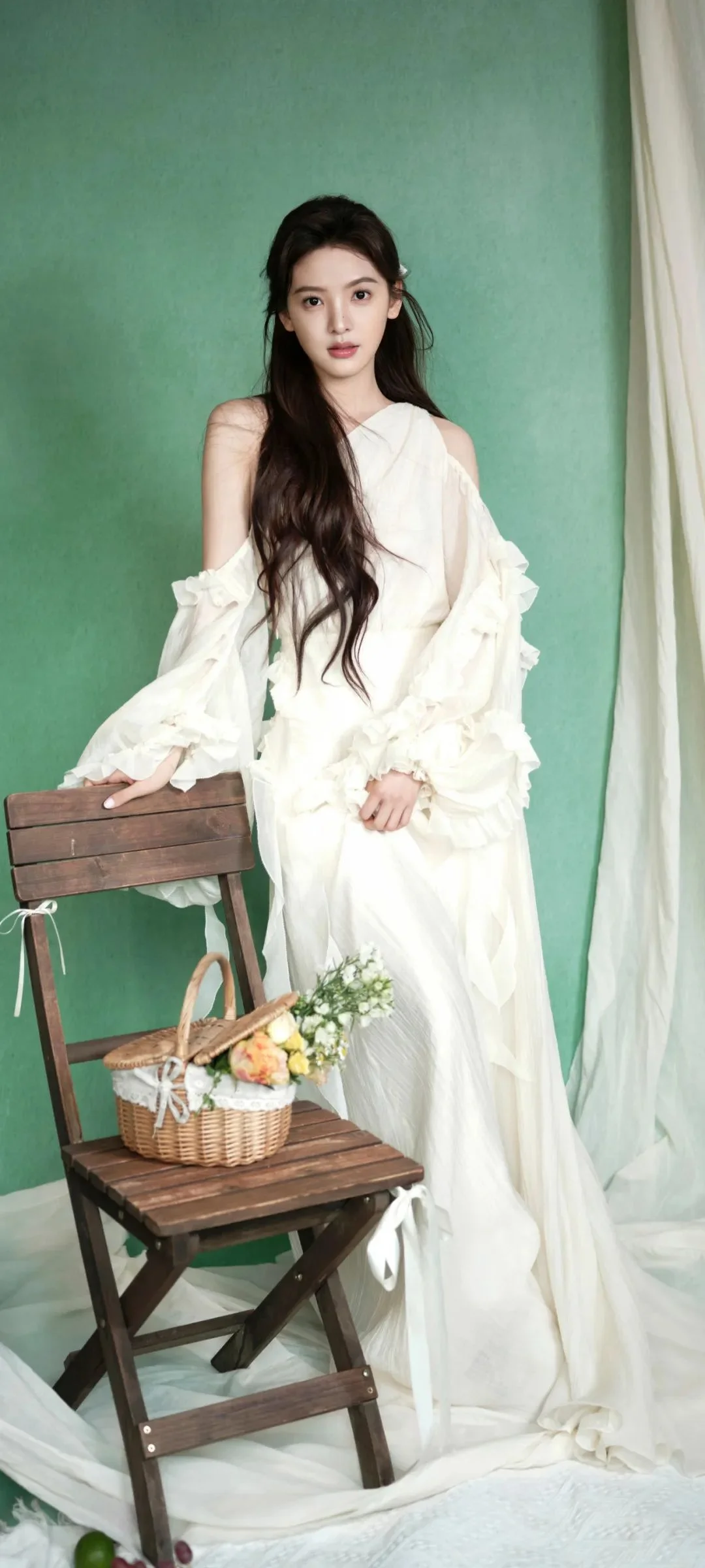 흰색 드레스를 입은 여성이 나무 의자와 꽃바구니 옆에서 초록색 배경 앞에 서 있다.