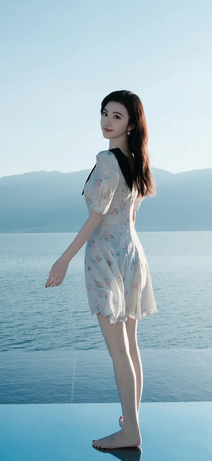 一名身穿浅色连衣裙的女子站在湖边，远处是群山与开阔的水面。