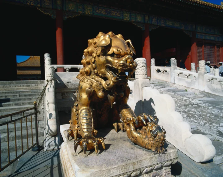 一尊鎏金石狮坐落在带有红柱和彩绘屋檐的古建筑石台前。