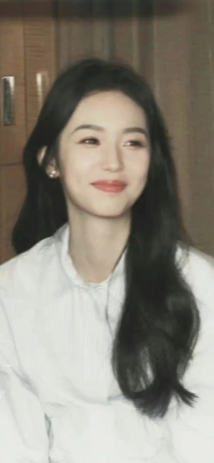 一名长发女性身穿白色上衣，在室内木质背景前微笑看向一侧。