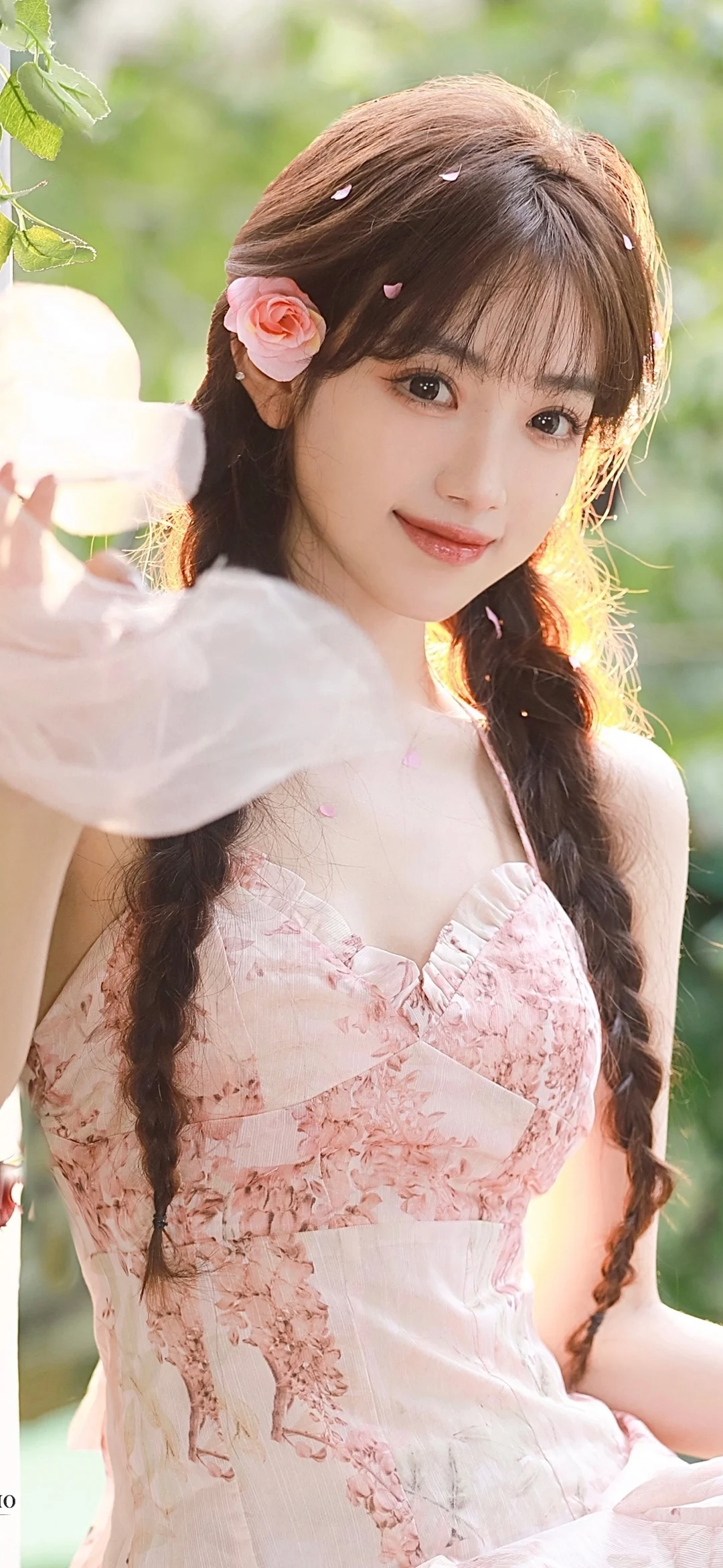 三つ編みと花の髪飾りをつけた女性が、木漏れ日の緑を背景にピンクの花柄ワンピース姿で微笑んでいる。