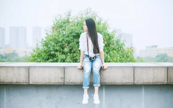 一名女子坐在屋顶边缘，身后是树木和朦胧的城市景观。