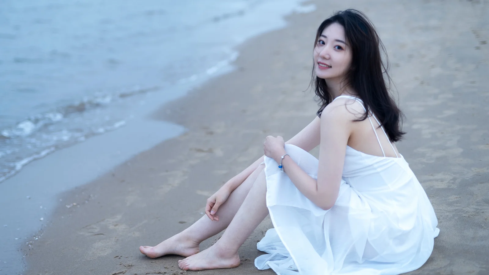 흰색 원피스를 입은 여성이 잔잔한 바다와 모래사장을 배경으로 해변에 앉아 있습니다.