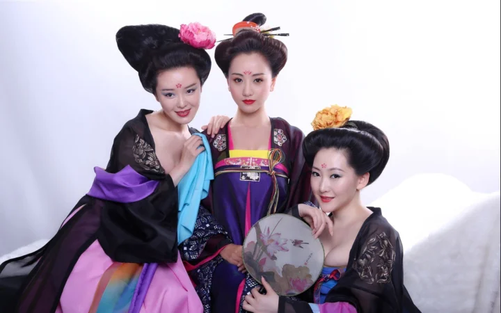 三名成年女子身穿传统服饰，在白色背景前合影，其中一人手持绘花团扇。