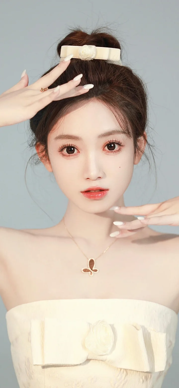 一位盘发女性身穿白色礼服，在浅灰色背景前直视镜头，双手靠近脸部。