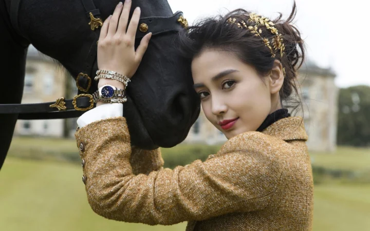 一名戴着金色发饰、穿着棕色外套的女士站在黑马上方，背景是户外草地和建筑。