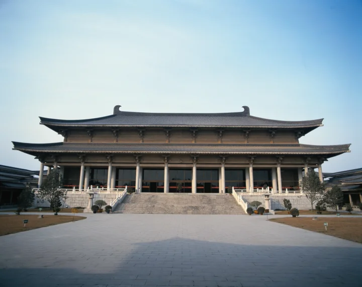 一座大型传统寺院建筑坐落在开阔的石板庭院前，背景是晴朗的浅蓝天空。