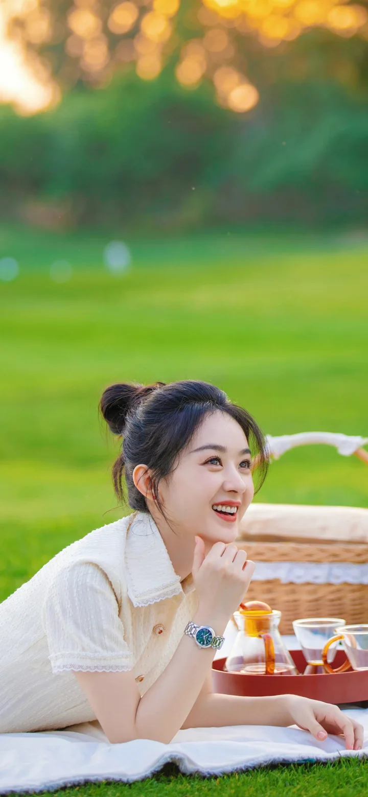 一名女子坐在绿草地上的野餐垫旁，面带笑容，身后是柔和虚化的户外景色。