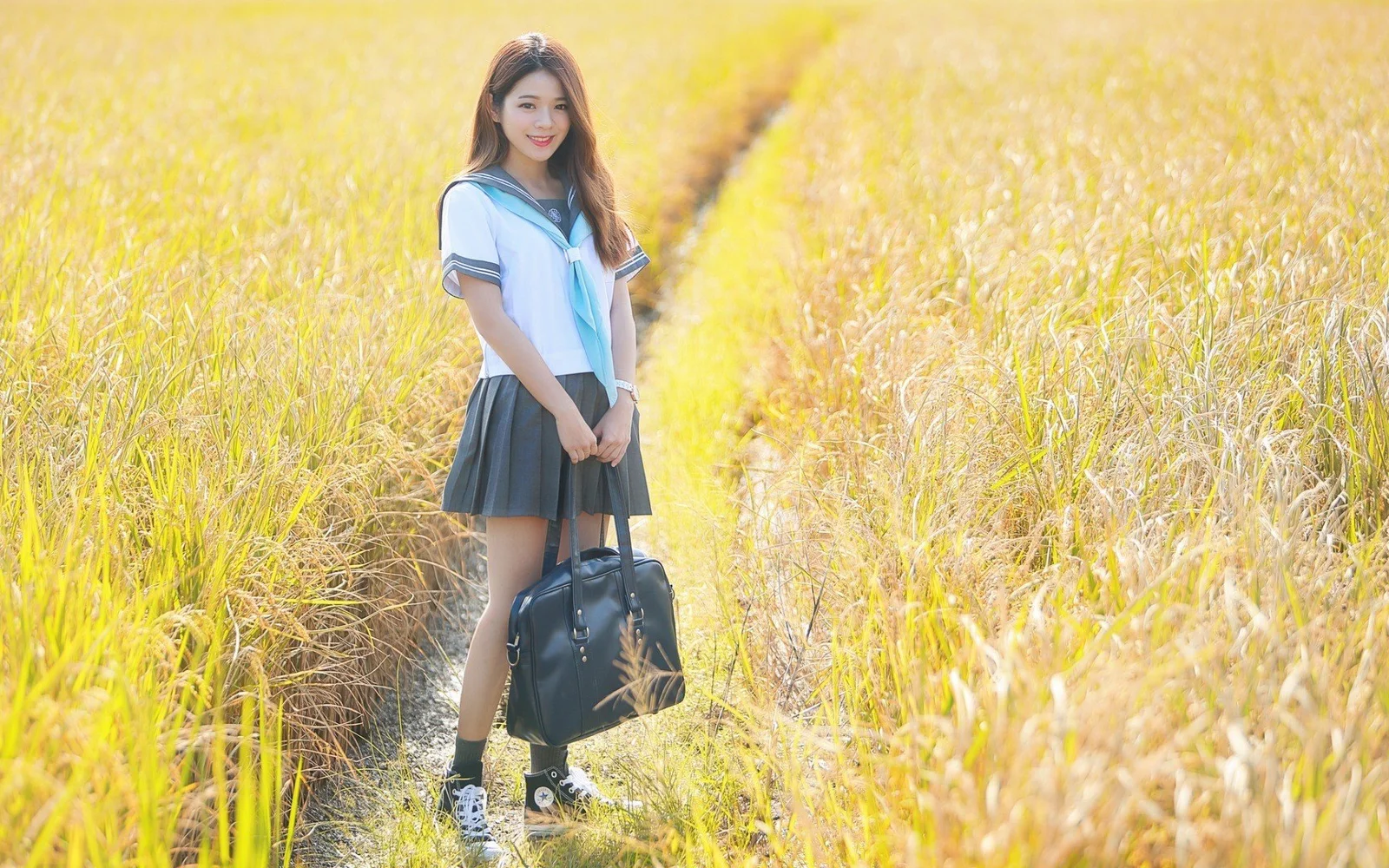 一名年轻女子站在金色草地间的小径上，手提黑色包袋。