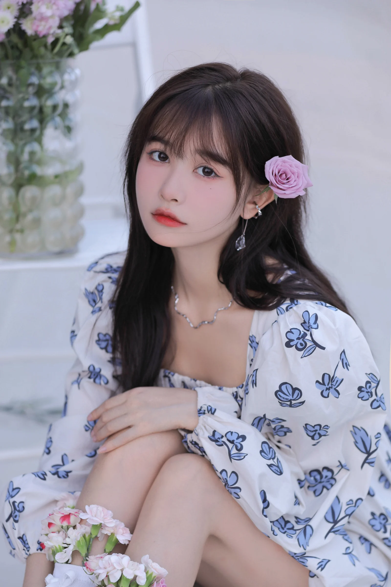 一位长发女性佩戴粉色玫瑰发饰，身穿蓝白花卉连衣裙坐在室内。