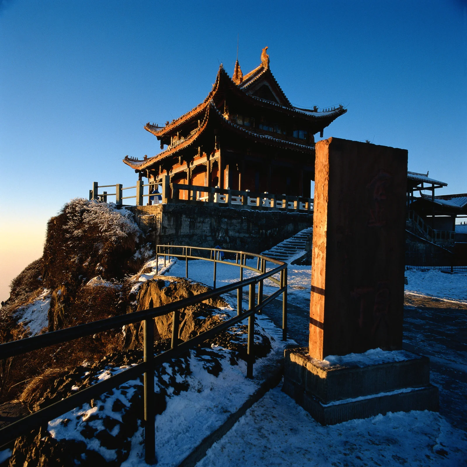 一座传统中式亭阁坐落在覆雪的悬崖边，蓝天下可见蜿蜒的栏杆和石阶。