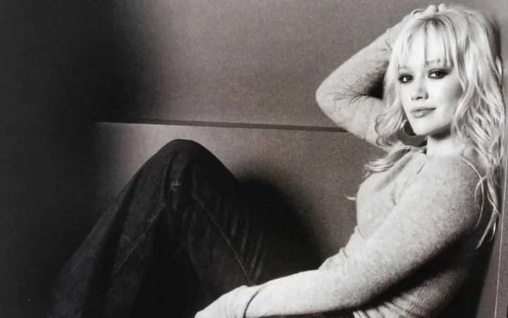 明るいトップスと濃い色のジーンズを着た金髪の女性が、室内で座る姿を写したモノクロポートレートです。