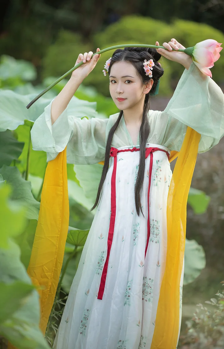 一位身穿浅色汉服的女性在荷塘边举起荷花枝，周围是荷叶与绿色植被。