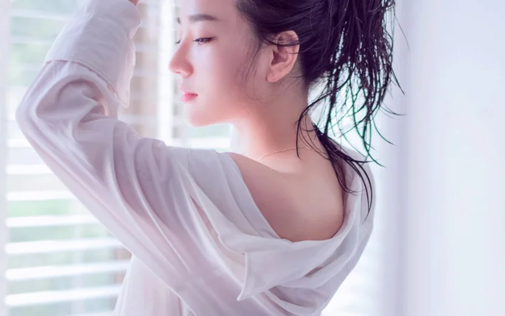 一位成年女性身穿宽松白衣站在窗边，侧脸与湿润的头发映入柔和的室内光线中。