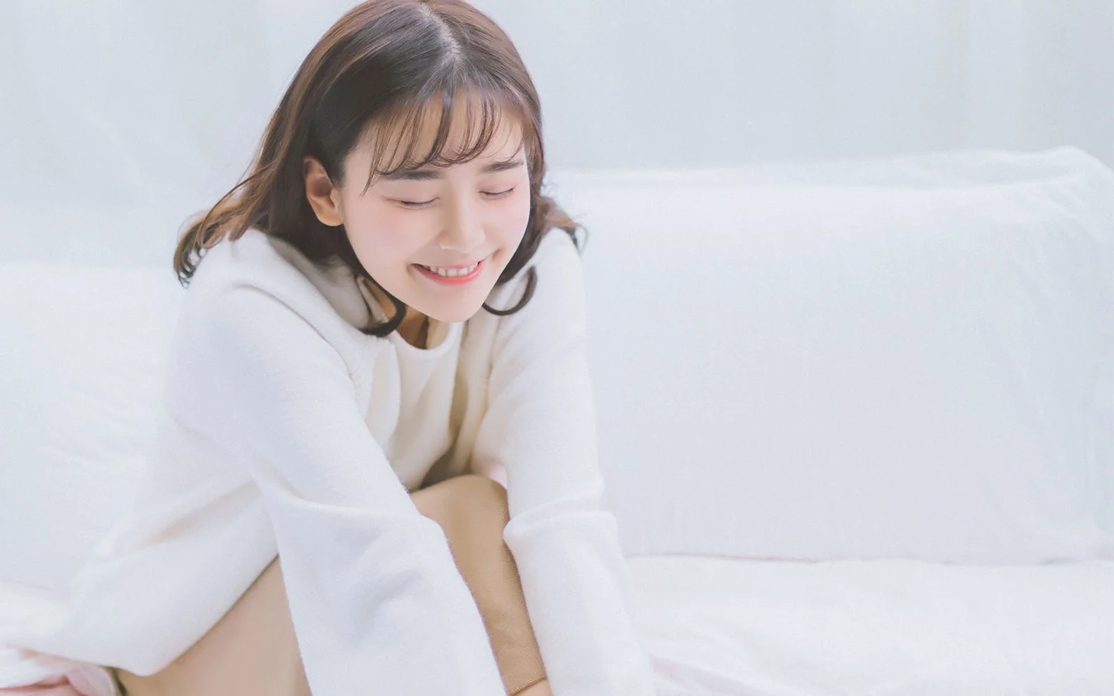 흰색 스웨터를 입은 성인 여성이 흰 침대에 앉아 미소 짓고 있다.