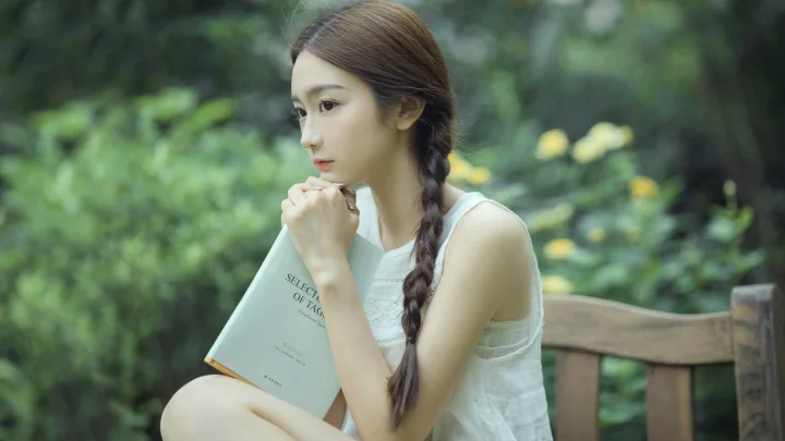 一名编着长辫的女性坐在户外长椅上，手持一本书望向一侧。