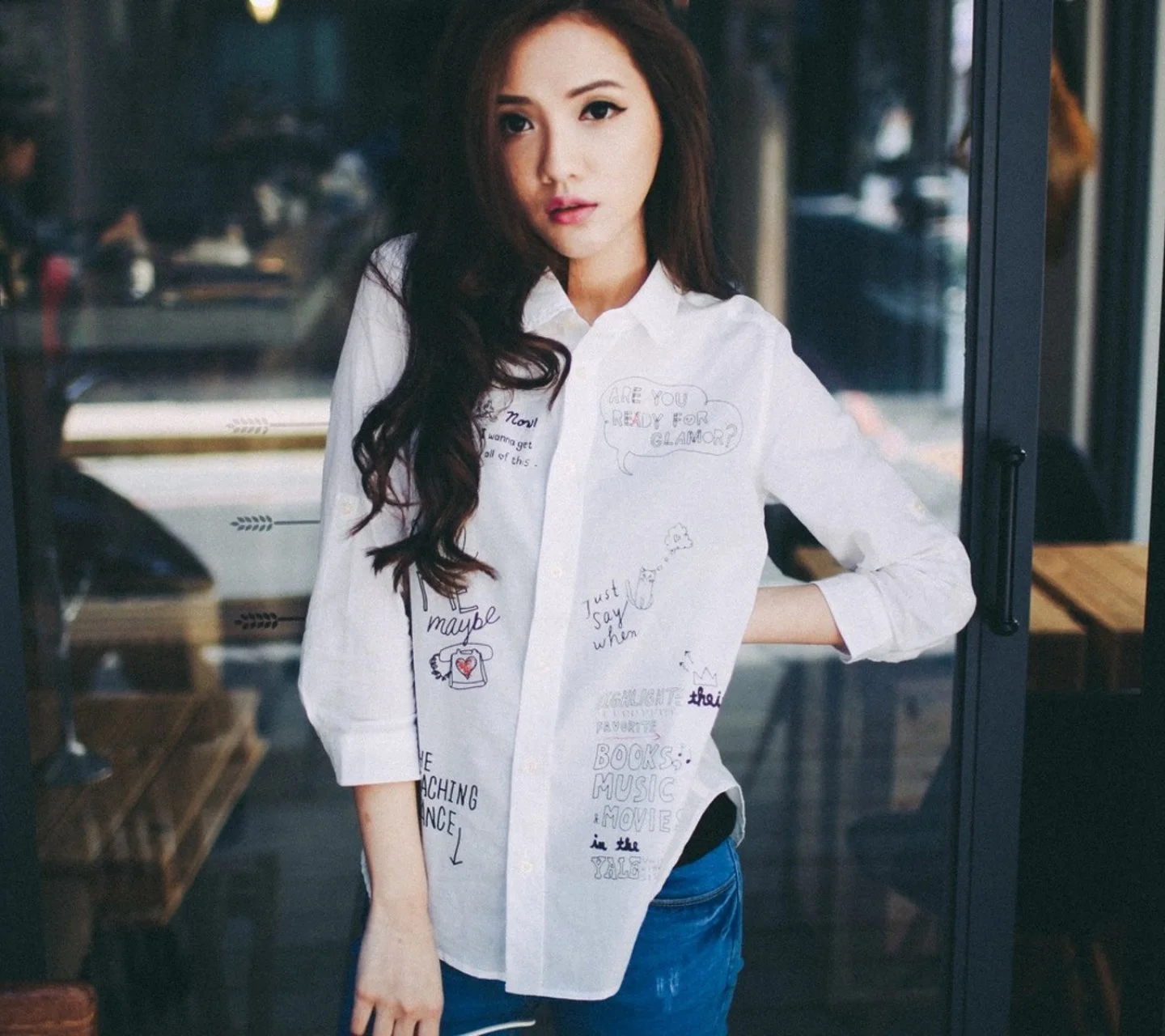 一名长发女性穿着带有文字图案的白衬衫站在室内窗边。