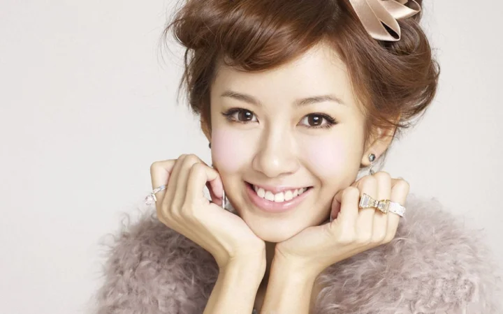 一位面带笑容的成年女性以双手托腮，身穿浅色绒毛服饰并佩戴发饰和饰品。