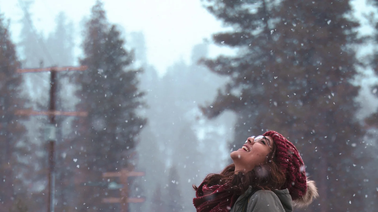 一名戴红色针织帽和围巾的女性站在飘雪的森林中仰望天空。
