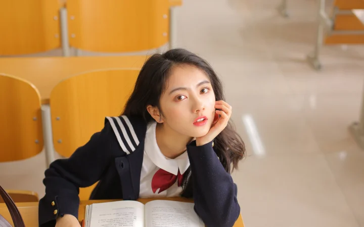 一名穿深色制服的年轻女性坐在教室课桌前，手边放着一本打开的书。