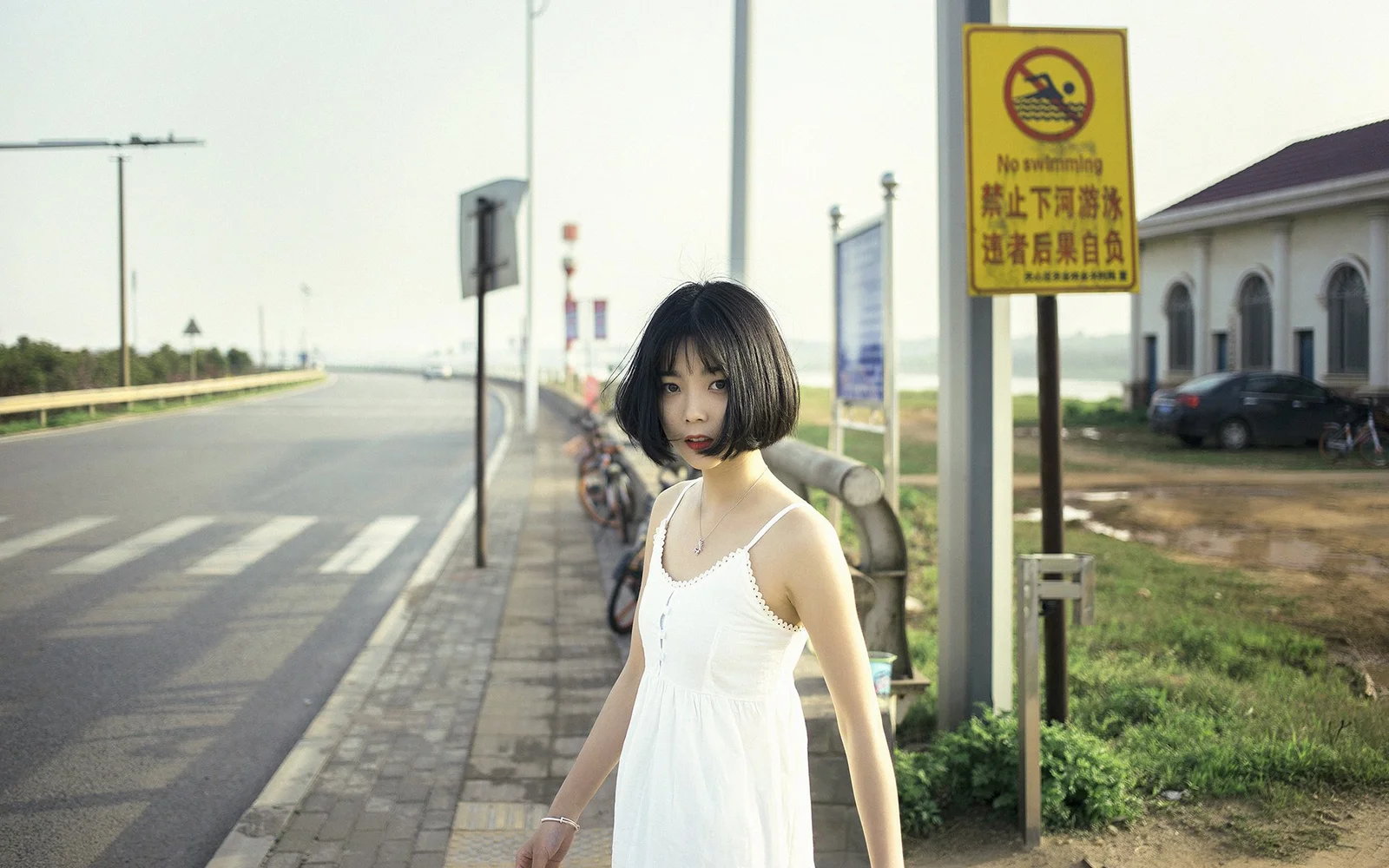 一名穿白色连衣裙的女子站在路边的人行道旁，身后可见道路、车辆和建筑。