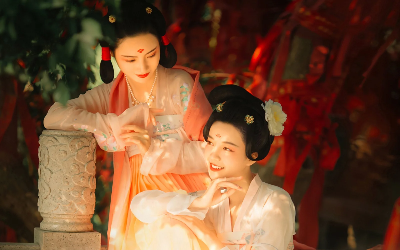 两名身穿传统服饰的女性在红色树木背景前摆姿势。