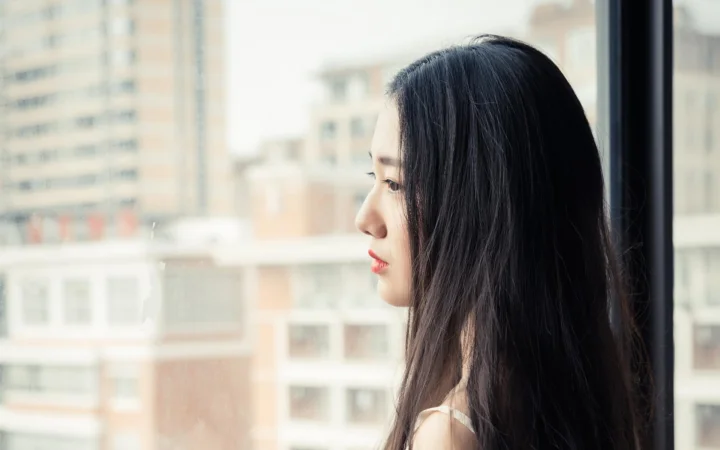 一名成年女性站在窗边，以侧脸望向窗外，背景是虚化的城市建筑。