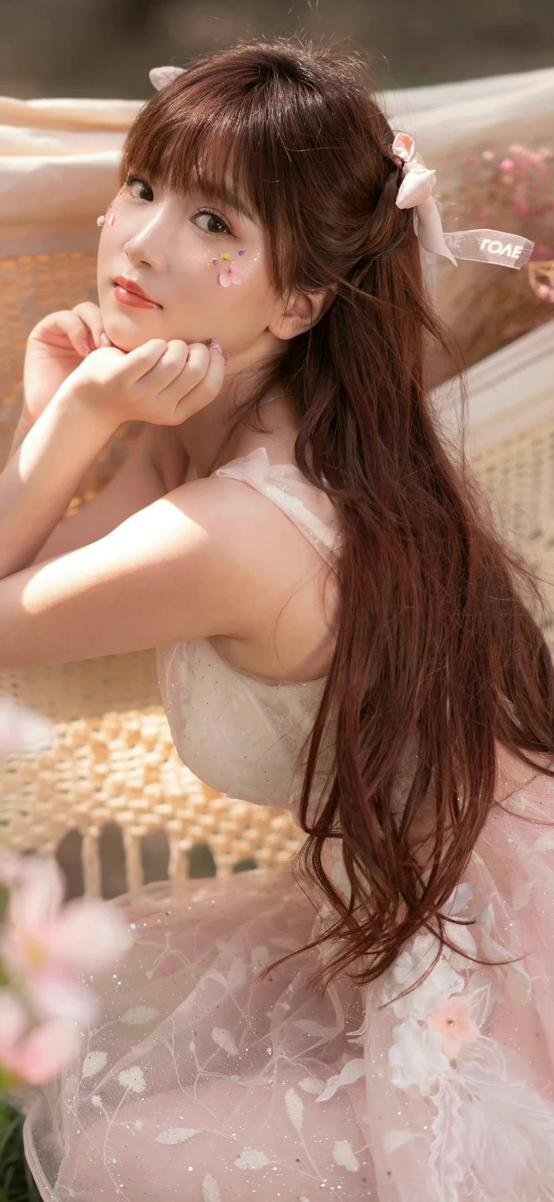 一名长发女性身穿浅色连衣裙坐在室内，双手托腮看向镜头。