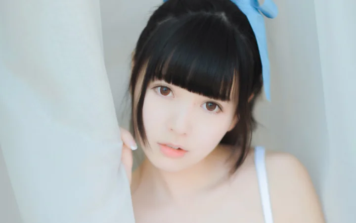 黒髪の前髪をそろえた女性が、明るく柔らかな背景の前で近距離からカメラを見つめている。