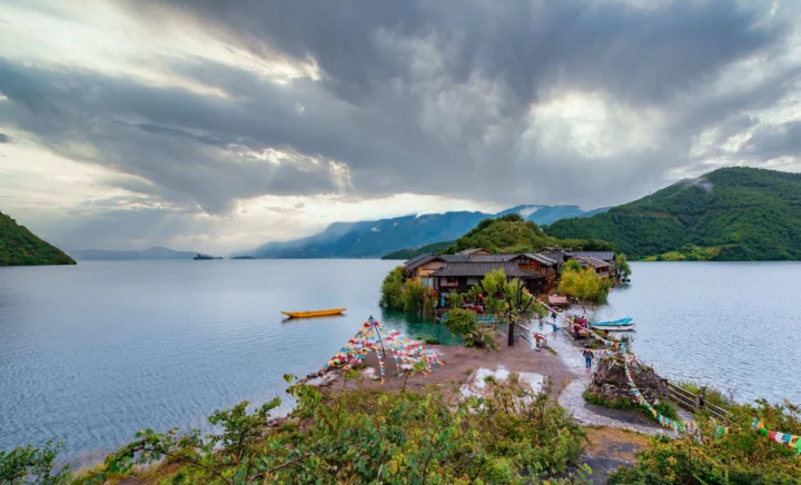 湖水延伸至远处群山之间，岸边坐落着木质建筑，厚重云层覆盖天空。