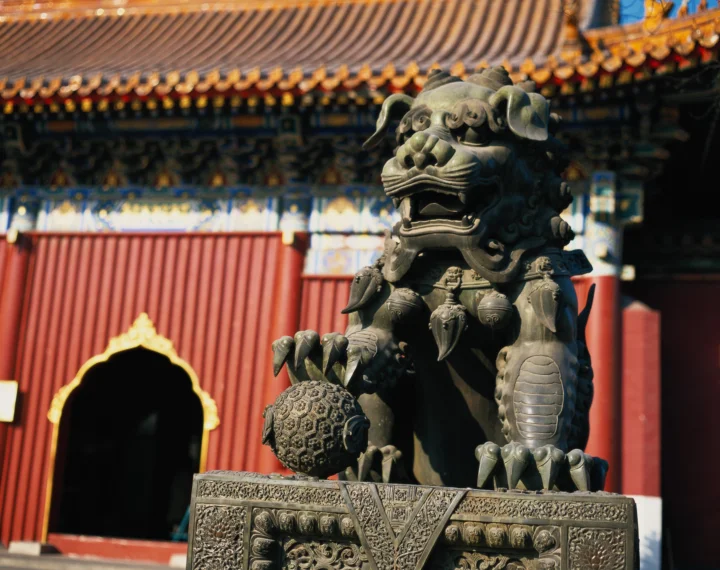 一尊雕刻精美的石狮矗立在中国传统宫殿建筑前，背景可见红墙、琉璃瓦和彩绘装饰。